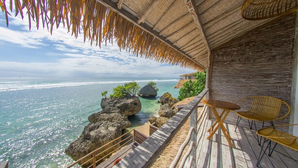 A wooden deck with a view of the ocean, a thatched roof, and two wicker chairs. - Le Cliff Bali (Pecatu)
