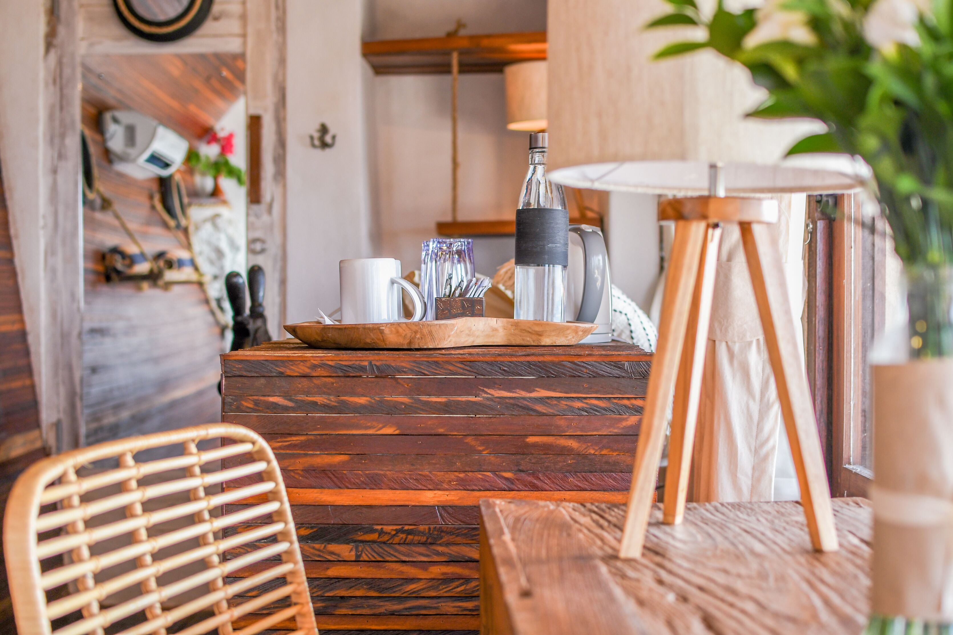 A wooden dining table with a white mug, a glass jar, and a metal container. There is a wicker chair in front of the table.