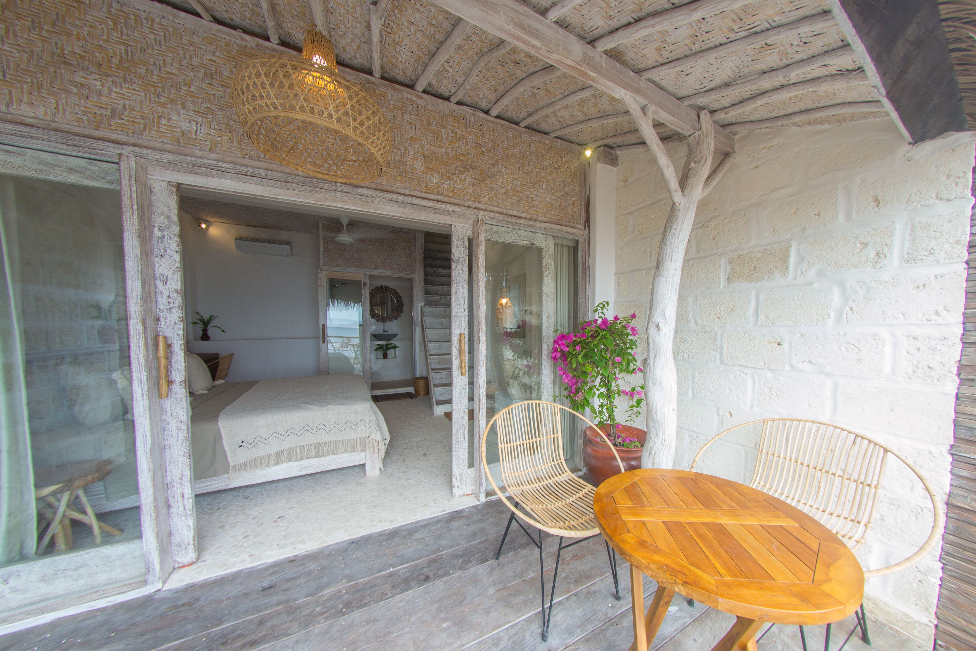 A room with a bed, a wooden table, two wicker chairs, a potted plant, and a glass door leading to another room.
