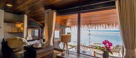 A room with a wooden ceiling, a wooden desk with a lamp, a chair, a bed with pillows, and a window with a view of the sea.