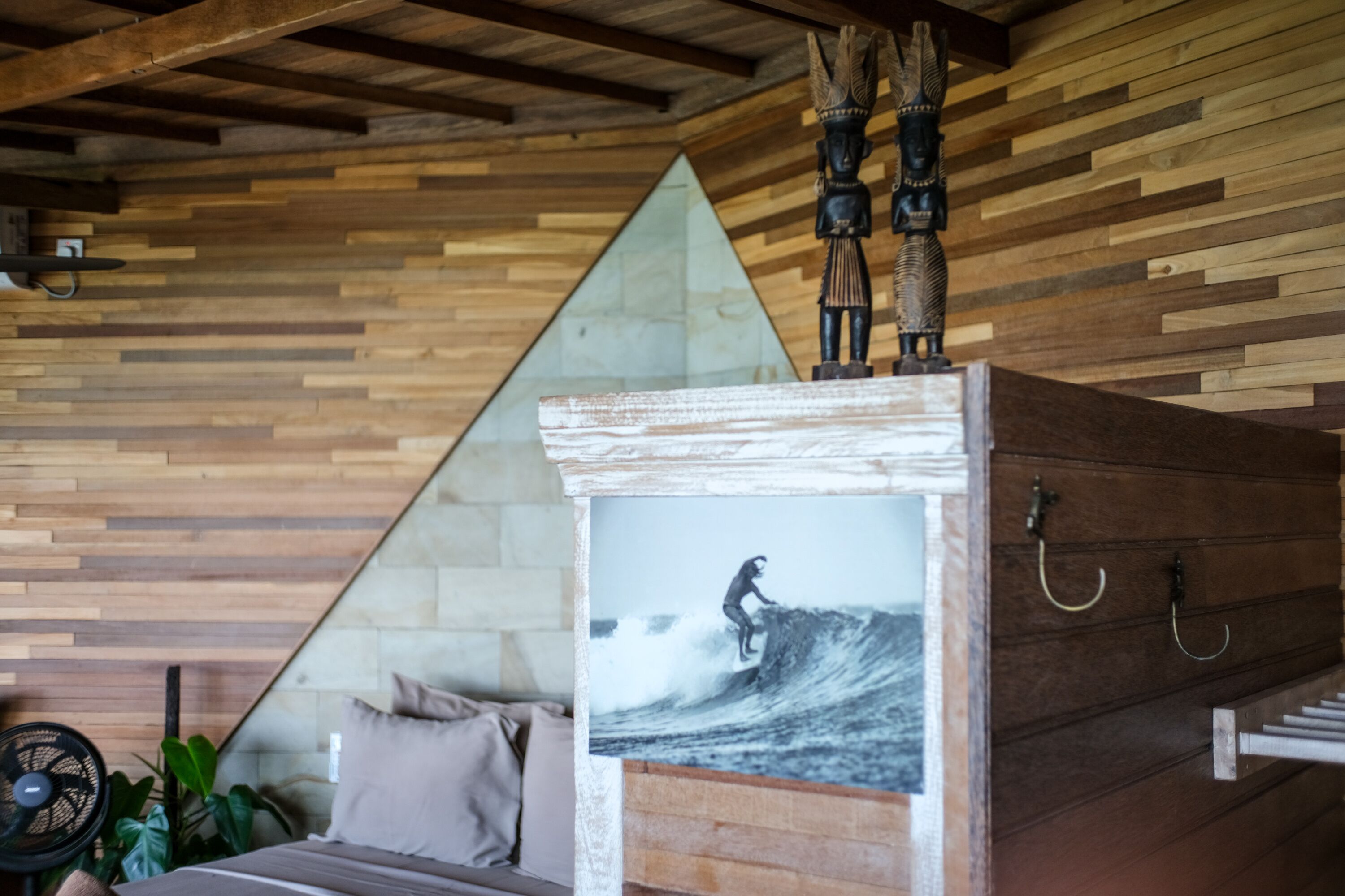 Ein Zimmer mit einer Wand aus Holzlatten, einem Bett mit Kissen, einem Ventilator und einem gerahmten Bild von einem Surfer.
