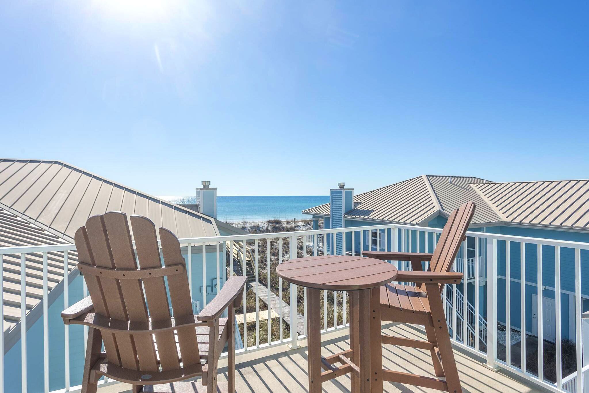 A wooden deck with two Adirondack chairs and a small table, overlooking a beach and ocean.
