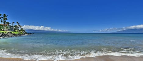 Une plage à l’eau d’un bleu limpide, bordée d’un rivage sablonneux, avec une chaîne de montagnes qui se dessine à l’horizon.
