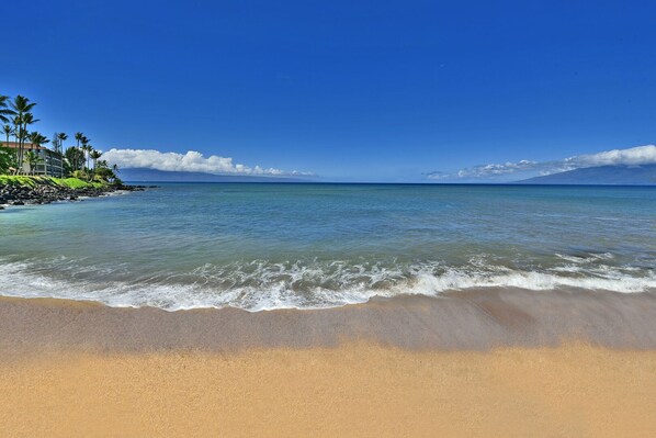 A beach with clear blue water, a sandy shore, and a distant mountain range.