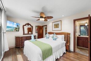 A hotel room with a bed, a wooden headboard, a ceiling fan, a large mirror, and a painting on the wall.