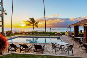 A pool area with lounge chairs and a barbecue grill, overlooking the ocean at sunset.