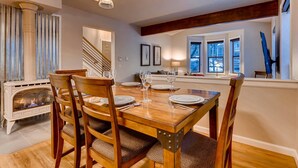 Un coin repas est aménagé avec une table en bois, dressée pour quatre personnes, et entourée de chaises dotées d’assises rembourrées. Une cheminée est visible sur la gauche, et un escalier mène à l’étage supérieur.