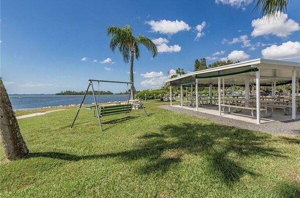 A park with a swing set, picnic tables, and a covered seating area by a lake.