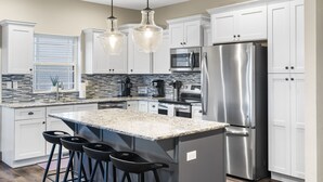 Una cocina moderna con gabinetes blancos, electrodomésticos de acero inoxidable, encimera de granito e iluminación colgante.