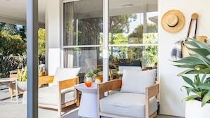 Un área de estar moderna al aire libre con sillas de madera y una mesa pequeña, ventanas amplias que ofrecen vistas a la vegetación y un sombrero de paja en la pared.