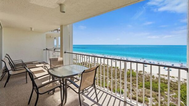 A balcony with a glass table and chairs, overlooking a beach with clear blue water and a rocky shoreline.