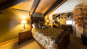 A cozy bedroom with a large bed, two bedside tables with lamps, a wooden ceiling, and a mirror reflecting the room. - Emmons Condo | 2 Bedroom plus Loft | Steps to Lifts | Shared Hot Tub | Parking (Mt. Crested Butte)