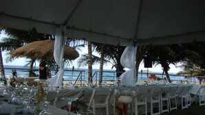 A tented dining area with tables set for a meal, overlooking a beach with palm trees and the ocean.