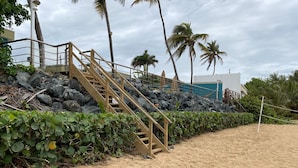 Une plage de sable avec un escalier en bois menant à un bâtiment, le tout entouré de palmiers et d’une haie.