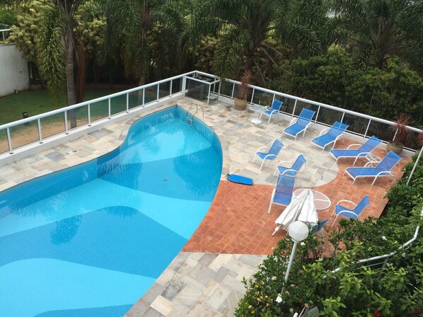 A swimming pool with a tiled deck, surrounded by blue lounge chairs and a white umbrella.