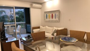 Ein modernes Wohnzimmer mit einem Glastisch, Korbstühlen, einer Couch und einem Balkon mit Blick auf Gebäude und Bäume.