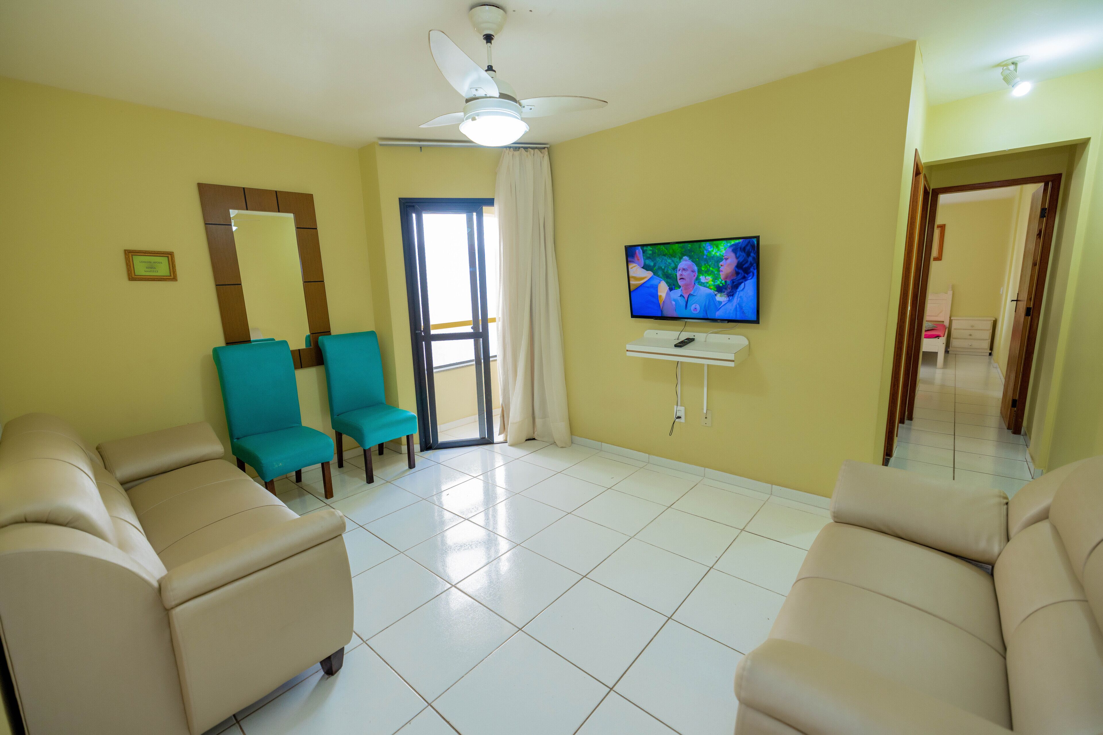 A living room with beige sofas, teal chairs, a television, and a ceiling fan.