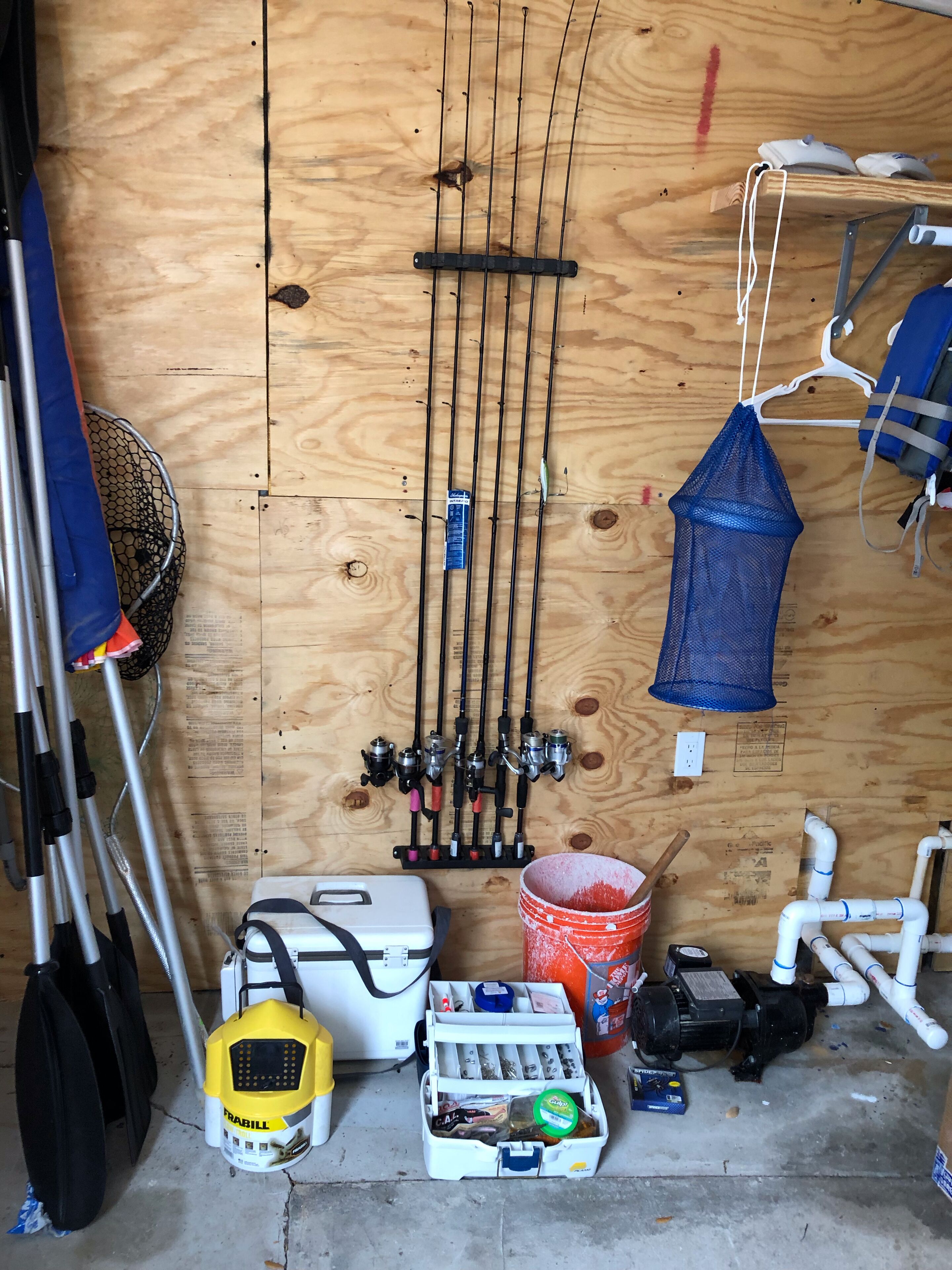 A wooden wall with fishing rods, a blue mesh bag, a white toolbox, a red bucket, and PVC pipes.