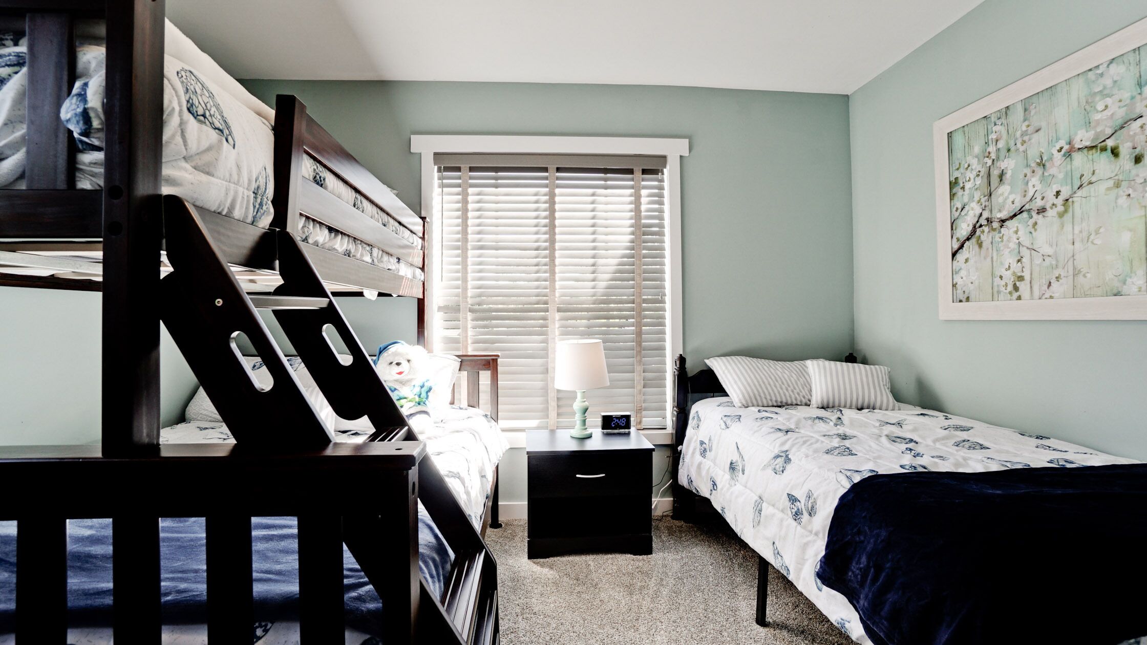 A bedroom with bunk beds, a bedside table with a lamp, a clock, and a window with blinds.