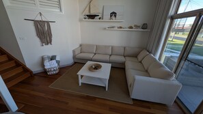 A living room with a beige sectional sofa, a white coffee table, and a wicker basket. There are shelves with decorative items and a view of the outdoors through a large window.