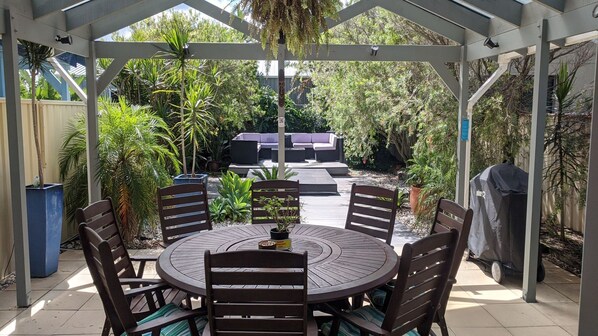 A covered patio with a round table and chairs, surrounded by lush greenery and a barbecue grill.