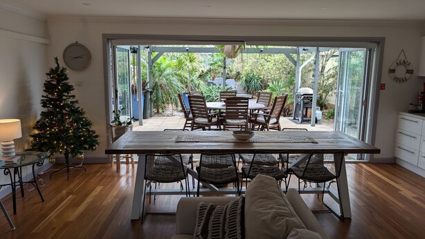 A dining area with a wooden table and chairs, a Christmas tree, and a view of the garden through glass doors. - Sunset on Birubi, 2/40 Ocean Ave - pet friendly, air con, water views, WIFI and outdoor spa (Anna Bay)