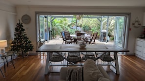 A dining area with a wooden table and chairs, a Christmas tree, and a view of the garden through glass doors.