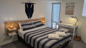 A bedroom with a bed, bedside tables, a desk, and a window with blinds. - Sunset on Birubi, 2/40 Ocean Ave - pet friendly, air con, water views, WIFI and outdoor spa (Anna Bay)