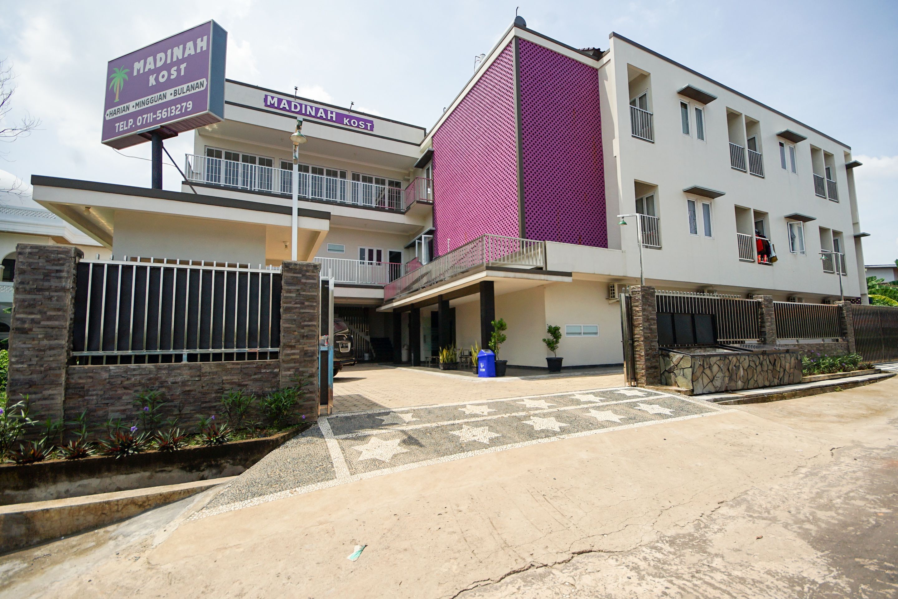 A modern building with a prominent purple facade, a gated entrance, and a signboard for 'Madinah Kost'.
