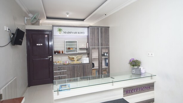A reception area with a glass counter, a dark wooden door, and a sign reading "Madinah Kost".