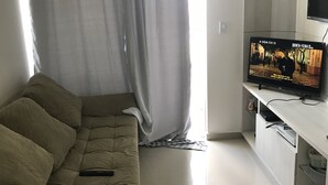 Ein Wohnzimmer mit einem beigen Sofa, dazu passend einem Hocker, einem Flachbildfernseher auf einem weißen Standfuß und einem Fenster mit Vorhängen.