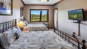 Ein Schlafzimmer mit zwei Betten, einem Deckenventilator, einem Fernseher und einem großen Fenster mit Blick auf Bäume und Berge.