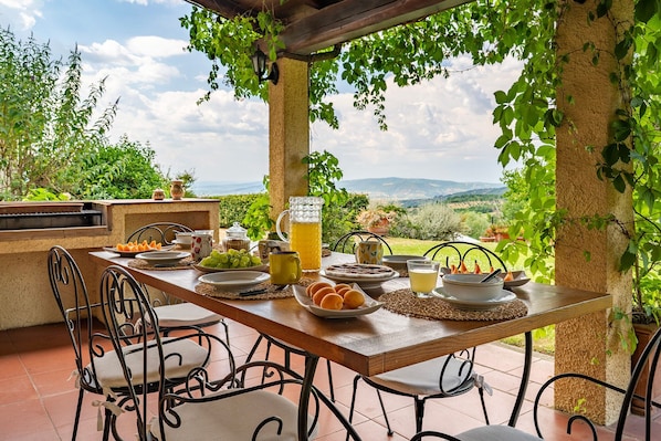An outdoor dining area with a view of the countryside, a table set with food and drinks, and a pergola structure.
