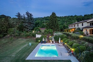 Una piscina rectangular con plataforma pavimentada, rodeada de césped cuidado y diferentes arbustos y árboles.