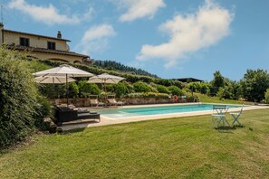 A swimming pool with lounge chairs and umbrellas, surrounded by greenery and a hill in the background.