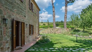 A stone building with a brick pathway leading to a grassy area with trees and a vineyard in the background.