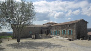 A stone building with a tiled roof, a gravel driveway, and a tree in the foreground.