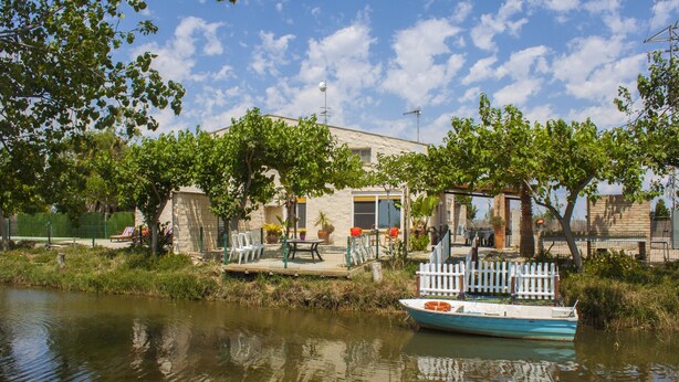 A houseboat docked at a calm river with a patio area, surrounded by greenery and a clear sky.