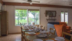 A dining room with a large table set for a meal, wooden chairs, a ceiling fan, and large windows with a view of greenery.