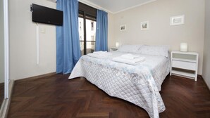 Een slaapkamer met een bed, een flatscreen televisie, een nachtkastje en een raam met gordijnen.