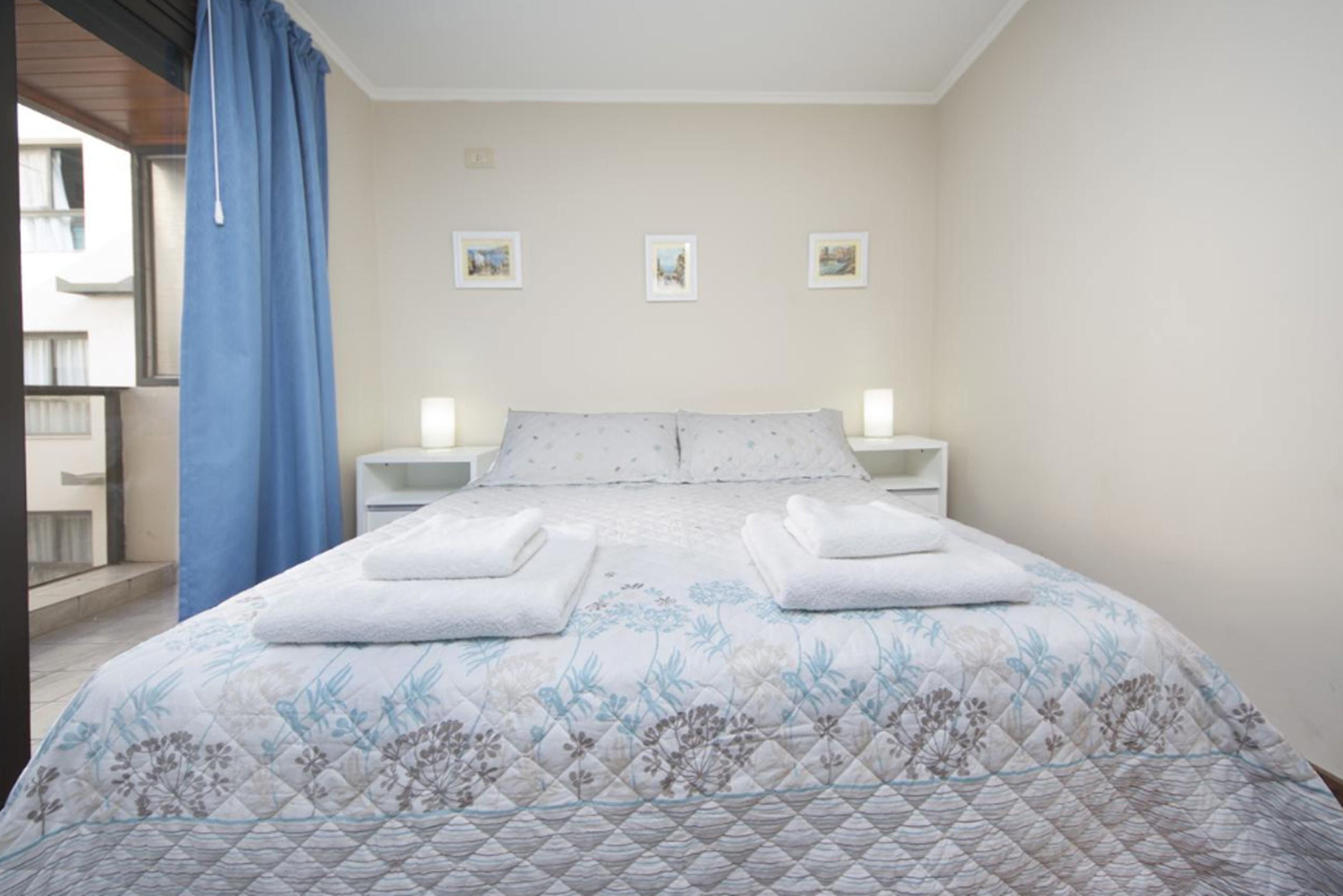 A hotel room with a bed, two bedside tables, a window with a blue curtain, and three small wall-mounted pictures.