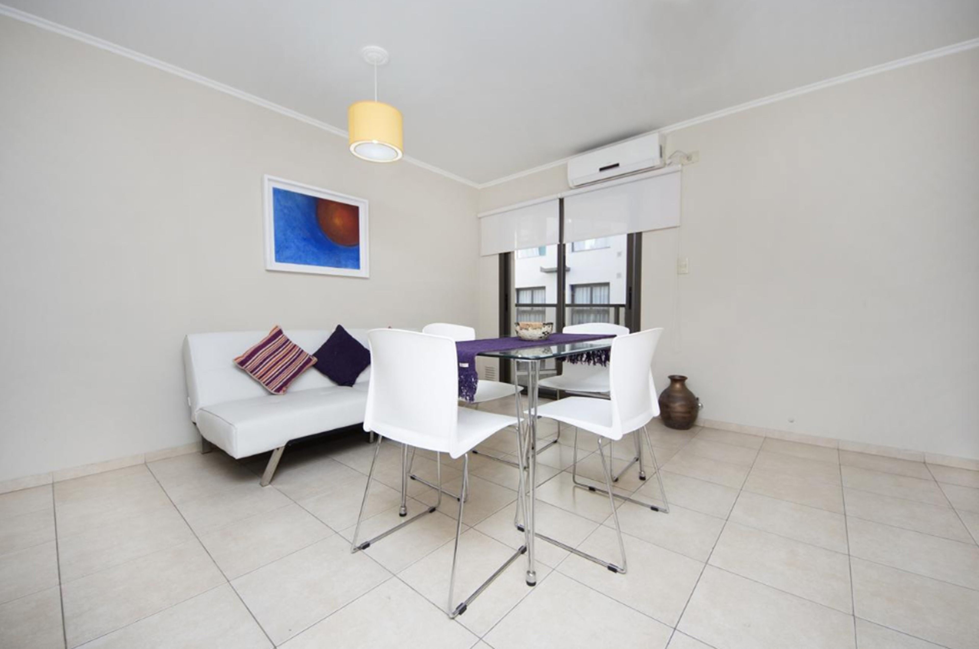 Um espaço de estar compacto com um sofá branco, uma mesa de jantar de vidro e uma luminária pendente amarela.