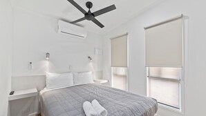 Une chambre moderne avec un lit, des murs blancs, un ventilateur de plafond, un appareil de climatisation, des tables de chevet et une fenêtre avec des stores.