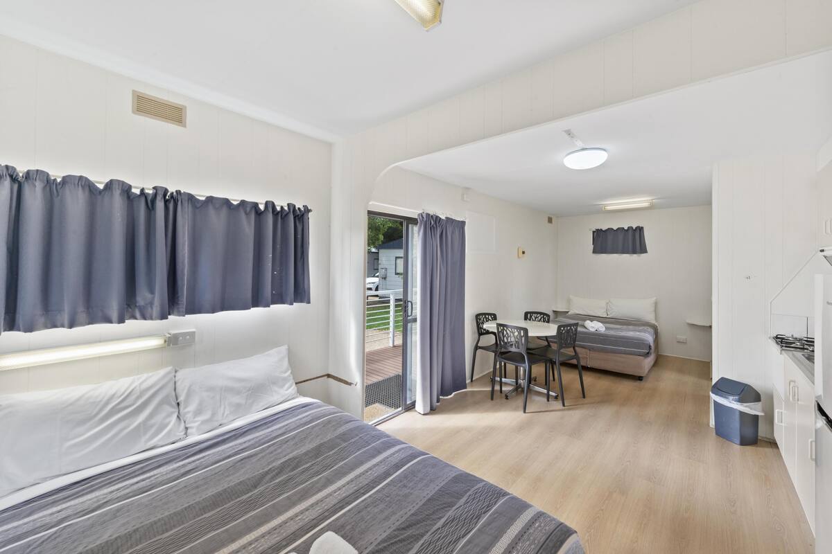 Ein Schlafzimmer mit Bett, Schreibtisch und Stuhl, einer Küchenzeile und Blick durch eine Tür.