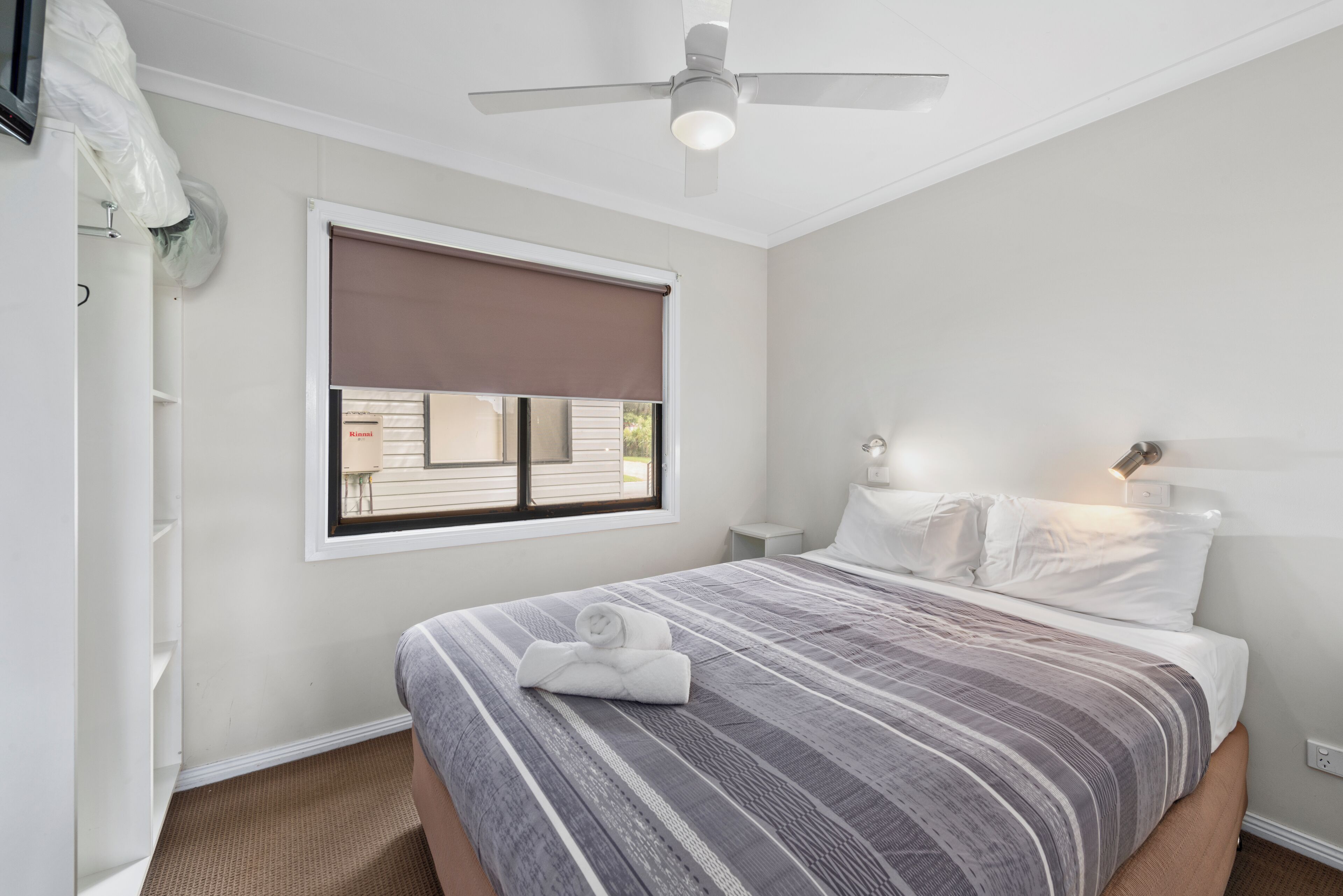 A bedroom with a bed, a ceiling fan, a window with a blind, and a nightstand with a lamp.