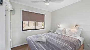 A bedroom with a bed, a ceiling fan, a window with a blind, and a nightstand with a lamp.