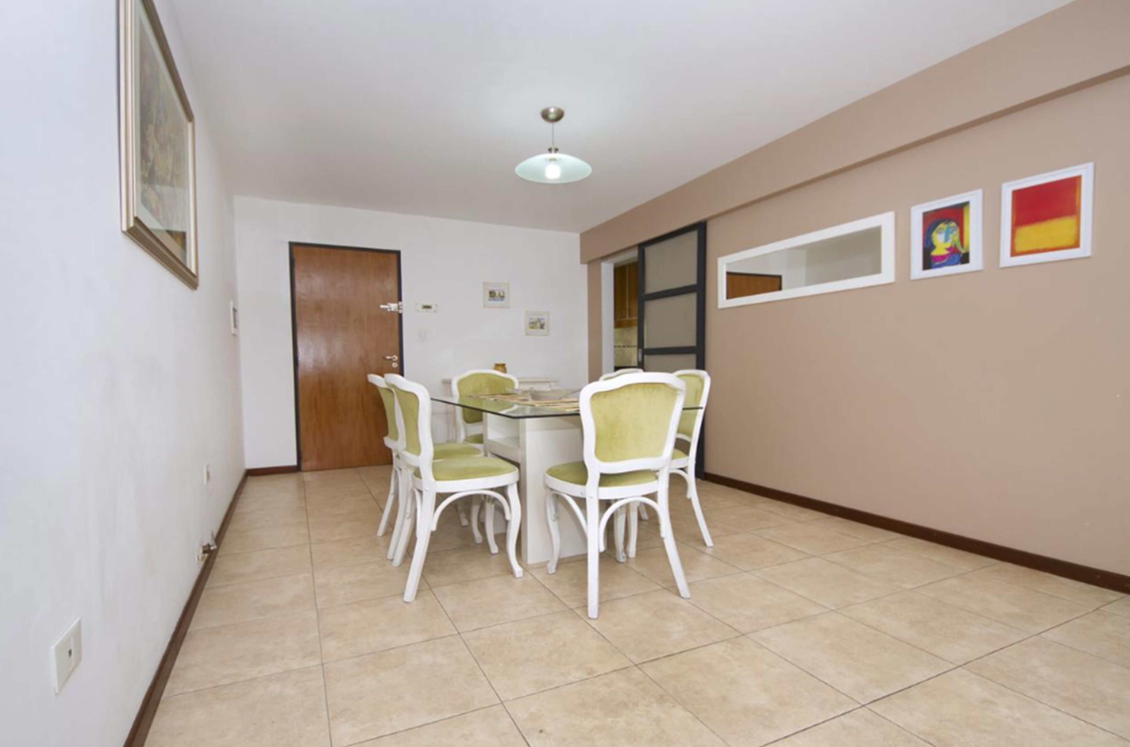 A dining area with a glass table and white chairs, a wooden door, and a beige wall with three framed pictures.