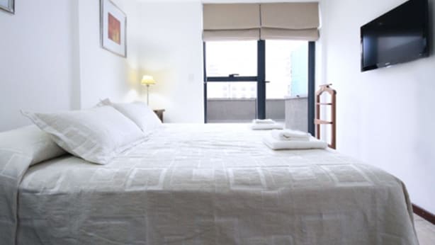 A hotel room with a bed, a TV mounted on the wall, and a window with a view of buildings. - Fragueiro Flat BU6 (Córdoba)