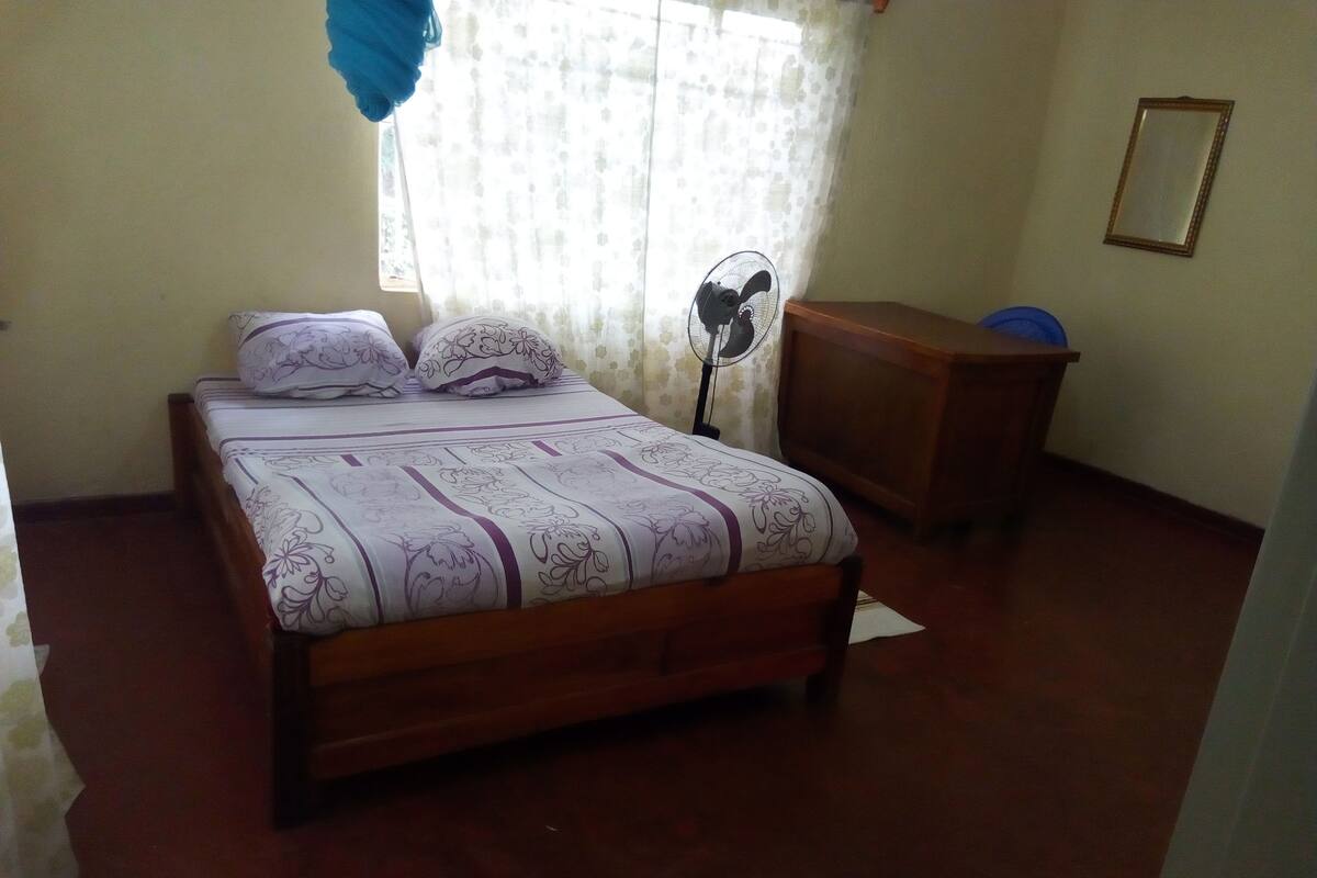 A bedroom with a wooden bed, a bedside fan, a wooden dresser, and a window with sheer curtains.