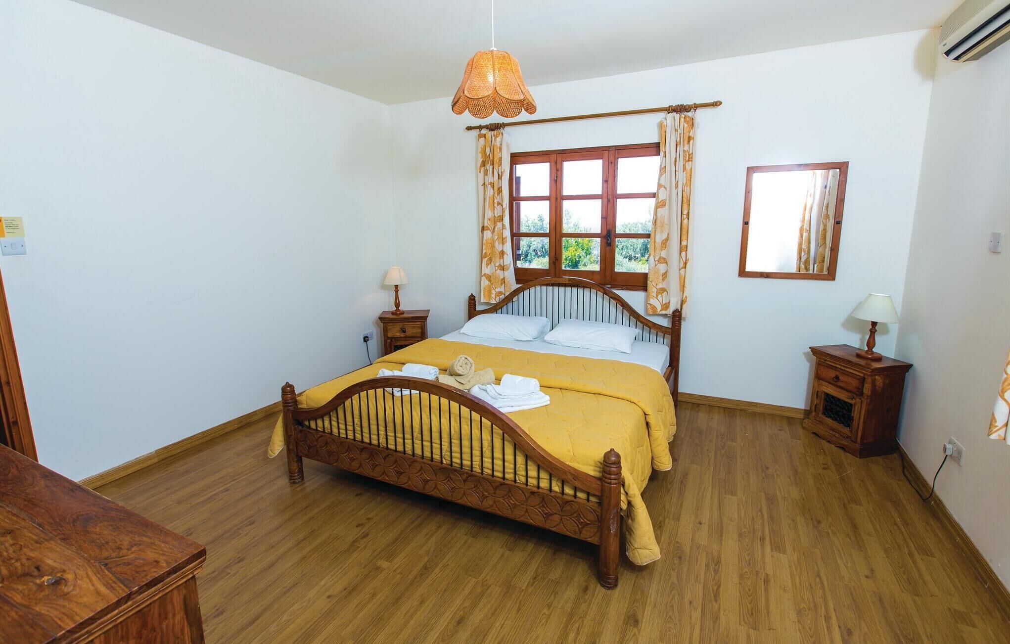 A neatly arranged bedroom with a wooden four-poster bed, a window with curtains, a bedside table with a lamp, and a mirror on the wall.